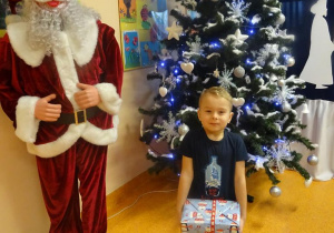 Uśmiechnięty chłopiec trzyma w ręku prezent, stoi obok figury Mikołaja.